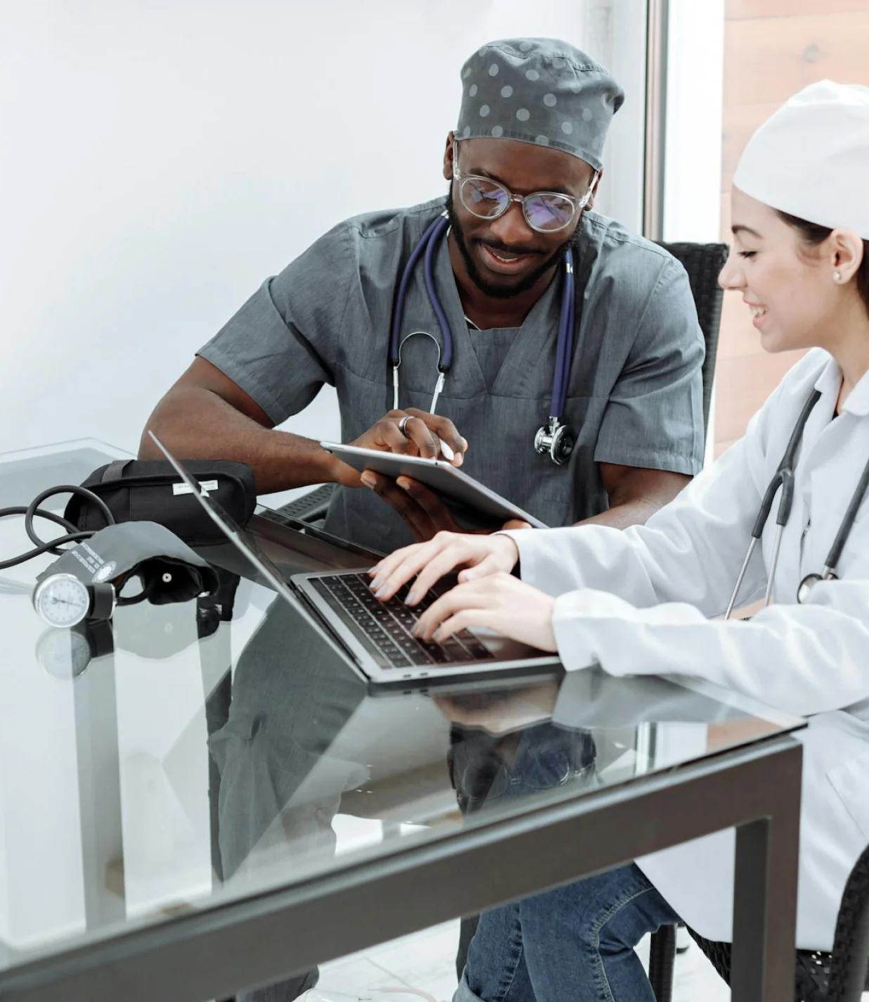 Two Nurses sitting at a table, each of them with a stethescope over their necks and going through records on a laptop